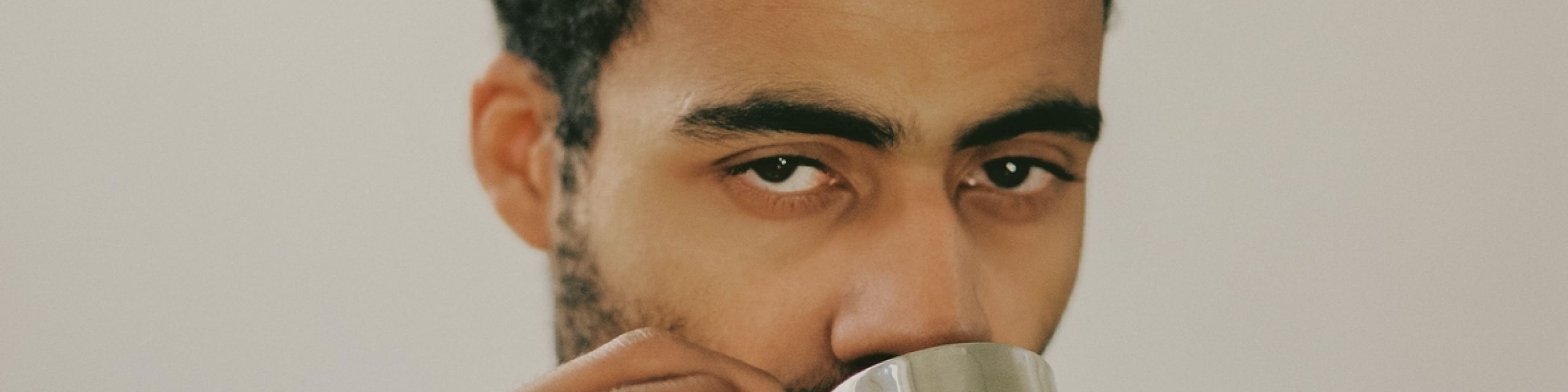Image alt: A person wearing a pearl necklace and two golden rings is looking at the camera full face and drinking a cup of espresso from a stainless steel cup