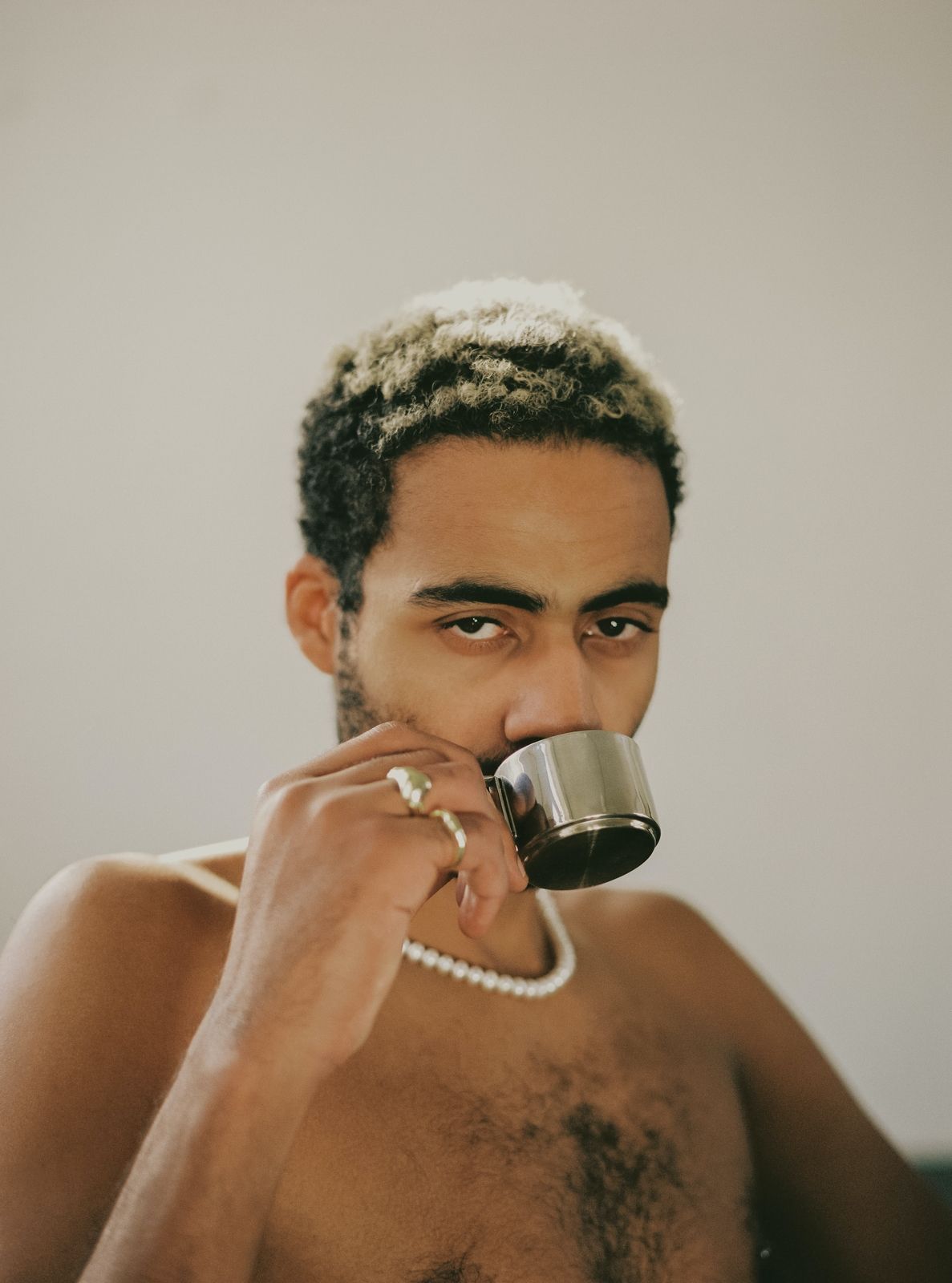 Image alt: A person wearing a pearl necklace and two golden rings is looking at the camera full face and drinking a cup of espresso from a stainless steel cup