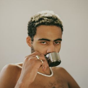 Image alt: A person wearing a pearl necklace and two golden rings is looking at the camera full face and drinking a cup of espresso from a stainless steel cup